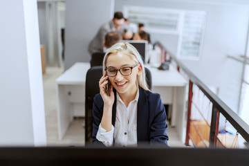 Wall Mural - Portrait of smiling Caucasian blonde female employee dressed in formal wear and with eyeglasses using computer while taking on the phone.