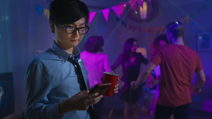 Wall Mural - At the Wild House Party: Confident Asian Man Uses Smartphone Instead of Dancing With Other People.