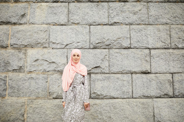 Wall Mural - Portrait of content beautiful Muslim lady in summer hijab and coat standing against old building