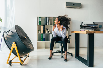 pretty african american businesswoman sitting on office chair in front of blowing electric fan