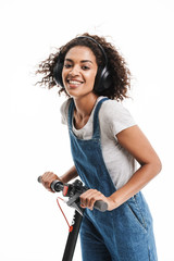 Wall Mural - Image of pretty african american woman dressed in denim overalls using headphones while riding on scooter