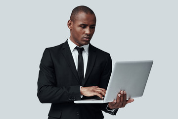 Ready to do business. Young African man in formalwear working using computer while standing against grey background