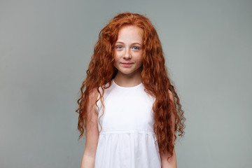 Wall Mural - Portrait of cute little girl with ginger hair and freckles, smiles and calming looks at the camera, stands over gray background.