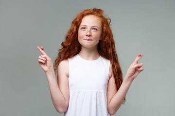 Wall Mural - Portrait of wishful nice little girl with ginger hair and freckles, calming smiles and looks up, crossed fingers and hopes for gud luck, stands over gray background.