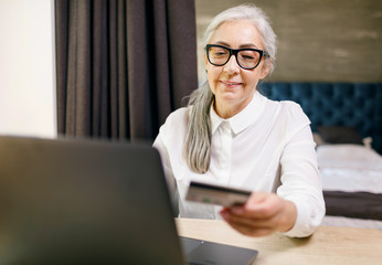 Cheerful aged long haired woman wearing glasses sitting with laptop and does online shopping in the internet with credit card