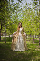  Beautiful girl in a gold dress against a background of green trees. A woman in a ball gown of the Rococo era.