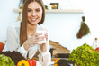 © rogerphoto - Young happy woman is holding white cup and looking at the camera while sitting at wooden table in the kitchen among green vegetables