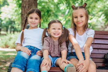 Three cute little girls smiling and sitting on the wooden bench in summer park