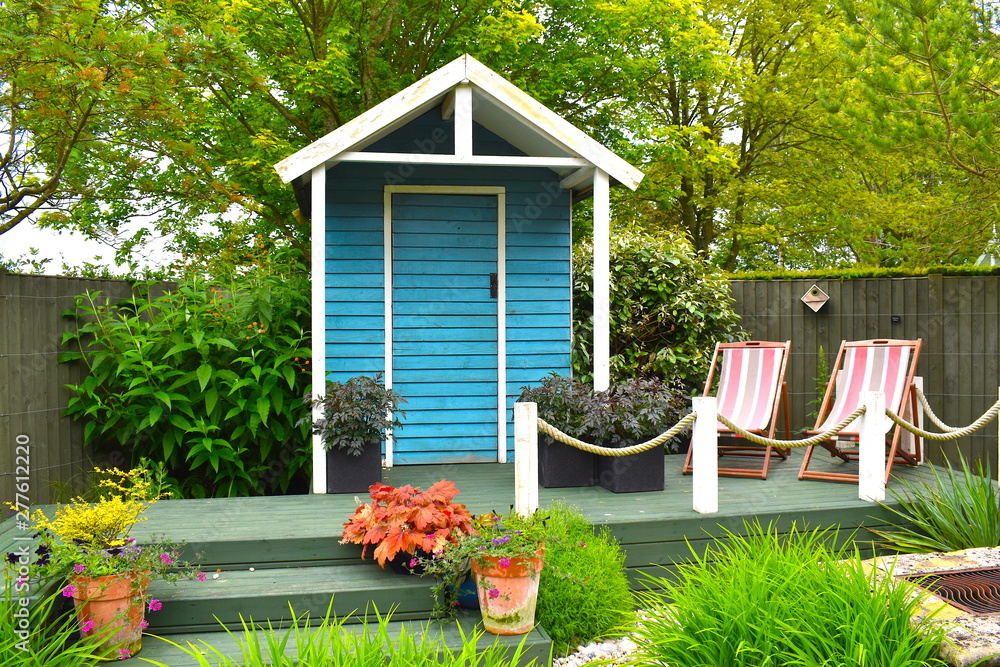 Wooden blue painted garden tools storage shed among green grass, potted plants and pink deckchairs in summer in the English countryside. Northamptonshire, UK - obrazy, fototapety, plakaty 
