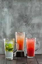Group Of Three Lemonade Jugs With Classic Lemonade, Wild Berry And Raspberry And Lychee Puree At Wooden Table Background.