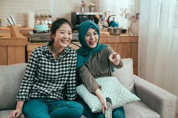 friendship people party entertainment concept. happy multi ethnic friends watching movie on tv at home. group of chinese and muslim women laughing and point at television screen while enjoy series.