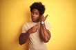© Krakenimages.com - American man with afro hair wearing striped t-shirt standing over isolated yellow background In hurry pointing to watch time, impatience, looking at the camera with relaxed expression