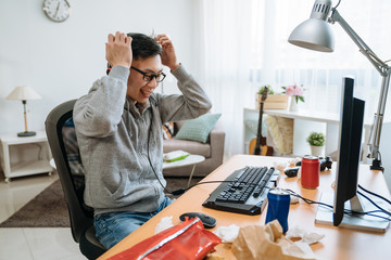 Wall Mural - asian guy playing online computer game sitting at desk. young man victory celebration raising hands indoors in bedroom. handsome male smiling cheerful while win on internet competition.