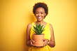 © Krakenimages.com - Young african american woman holding cactus pot over isolated yellow background with a happy face standing and smiling with a confident smile showing teeth