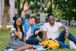 © Iryna - Happy positive group of university multiethnic students friends from different countries waving for you smiling while relaxing on the campus lawn during free time. Different culture friendship concept