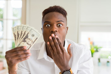 Poster - African american man holding twenty dollars bank notes cover mouth with hand shocked with shame for mistake, expression of fear, scared in silence, secret concept