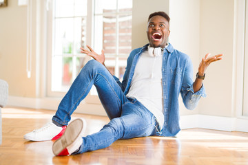 Poster - African american man wearing headphones listening to music sitting on the floor celebrating crazy and amazed for success with arms raised and open eyes screaming excited. Winner concept