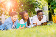 © Iryna - Front view of three happy multiethnic students lying, talking, joking together together on green lawn high school university campus. Education and Friendship Concept. Sun glare effect