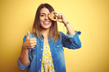 Young beautiful woman eating chocolate chips cookie over yellow background happy with big smile doing ok sign, thumb up with fingers, excellent sign