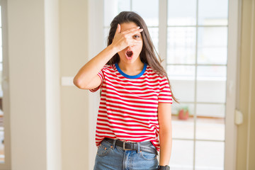 Poster - Young beautiful woman wearing casual t-shirt peeking in shock covering face and eyes with hand, looking through fingers with embarrassed expression.