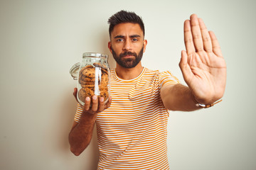 Poster - Young indian man holding jar of cookies standing over isolated white background with open hand doing stop sign with serious and confident expression, defense gesture