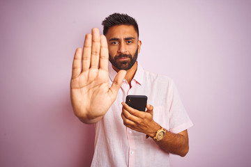 Young indian man using smartphone standing over isolated pink background with open hand doing stop sign with serious and confident expression, defense gesture