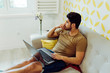 © Manuel Ruiz Alba/ADDICTIVE STOCK - Side view of young thoughtful man in casual outfit with laptop sitting on couch near table with fresh flowers