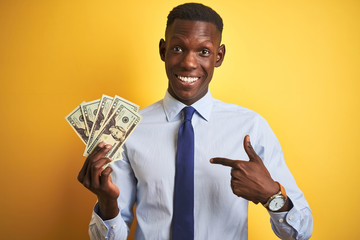 African american businessman holding dollars standing over isolated yellow background with surprise face pointing finger to himself