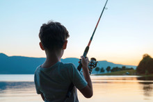 A Happy Boy Is Standing On The Shore Of A Beautiful Lake And Fishing With A Fishing Rod Against The Background Of A Bright Sunset In The Summer. Child On Summer Vacation With Family. Tourism. Camping