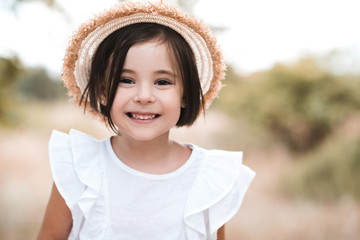 Wall Mural - Smiling kid girl 3-4 year old wearing straw hat and white stylish shirt outdoors. Looking at camera.