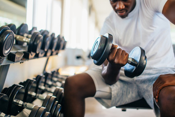 Wall Mural - Young African American man sitting and lifting a dumbbell close to the rack at gym. Male weight training person doing a biceps curl in fitness center