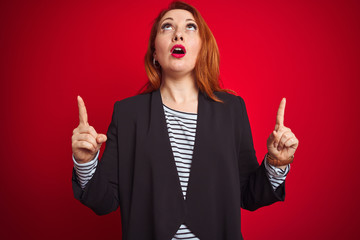 Sticker - Beautiful redhead business woman wearing elegant jacket over isolated red background amazed and surprised looking up and pointing with fingers and raised arms.