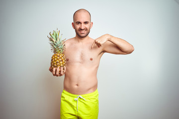 Wall Mural - Young man wearing summer hat and swimwear holding pineapple over isoalted background with surprise face pointing finger to himself