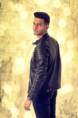 Wall Mural - Young handsome man standing in studio shot, on golden background, wearing black leather jacket and black jeans