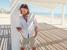 Handsome Bearded Man Using Smartphone And Looking Away While Leaning On Pillar Of White Gazebo On Sandy Beach