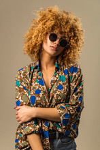 Attractive Ethnic Female With Blond Curly Hair Wearing Vintage Sunglasses And Ornamental Blouse While Standing Against Gray Background