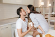 © Look! - Excited female model sits on table kissing man in white t-shirt, enjoying good morning. Indoor portrait of loving couple having fun during joint breakfast.