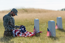 Sad Soldier In Camouflage Uniform And Cap Holding American Flag While Sitting In Graveyard