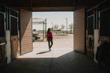 Back View Of Woman Walking Out Of Horse Stable On Ranch In Bright Sunshine