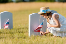 Selective Focus Of Adorable Kid In Straw Hat Sitting Near Headstone With American Flag In Graveyard