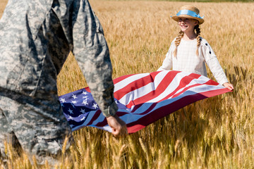 Wall Mural - cropped view of man in military uniform holding american flag with cute daughter in field