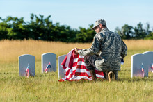 Man In Military Uniform And Cap Holding American Flag While Sitting In Graveyard
