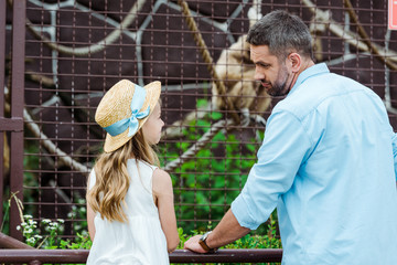 Wall Mural - kid in straw hat looking at upset father near cage with wild animal in zoo