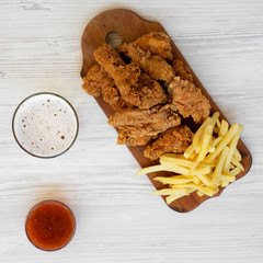 Wall Mural - Tasty fast food: fried chicken drumsticks, spicy wings, French fries and chicken fingers with sour-sweet sauce and glass of cold beer on a white wooden surface, top view. 