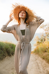 Wall Mural - Pretty cheerful happy smiling young woman in hat posing outdoors at the nature green grass near beach sea.