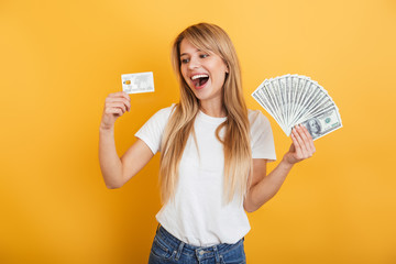 Poster - Optimistic positive young blonde woman jumping isolated over yellow wall background dressed in white casual t-shirt holding money and credit card.