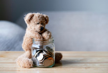 Teddy Bear Putting Hand Into The Jar Of British Coins On Wooden Table , Brown Bear Sitting Next To Clear Glass Jar With Pound Coins, Money Saving Concept