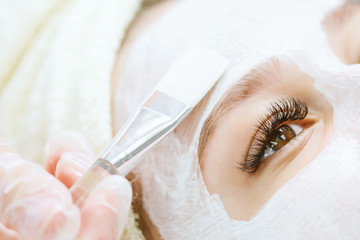 Wall Mural - Portrait of beautiful woman laying with towel on the head. Hands of cosmetology specialist applying cream facial mask using brush. Beauty, spa, cosmetology and skincare concept.