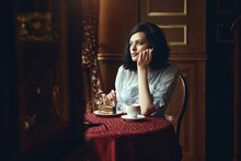 Portrait Of Young Beautiful Girl Sitting At The Table In The Cozy Coffee Shop And Looking At The Window Thoughtfully, Her Face Buried In Her Hands. Chocolate Cake And Cup Of Coffee On The Table