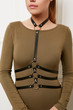 Closeup front shot of girl's chest. The woman is wearing olive dress with boat neck and leather chest harness with 3-rows of straps connected with rings. The woman is turning head on the white wall.